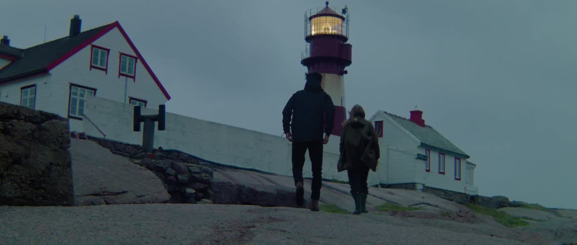 Man and woman with black coats and jeans walk up to lighthouse on cloudy day