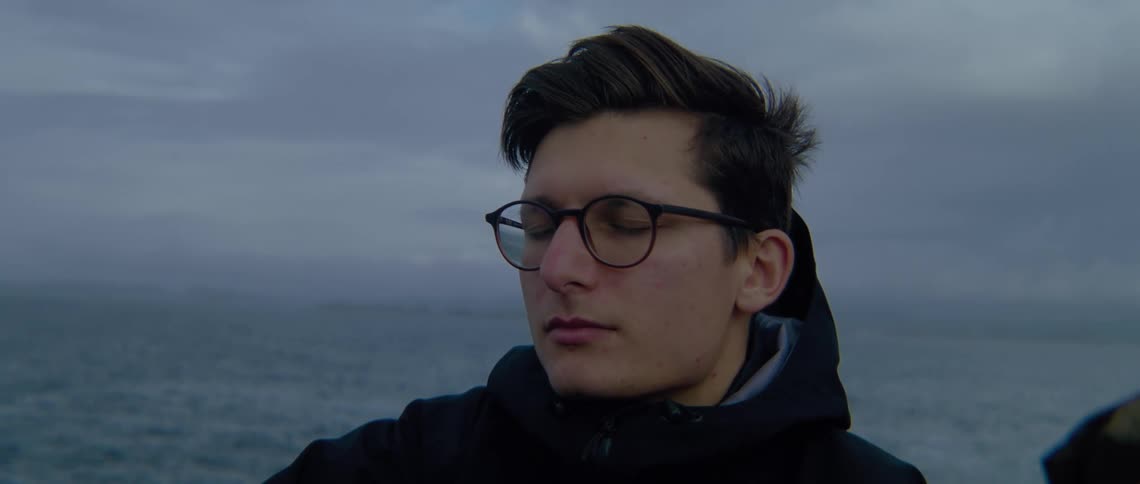 A man wearing glasses and a dark coat has his eyes closed relaxing with the ocean behind him