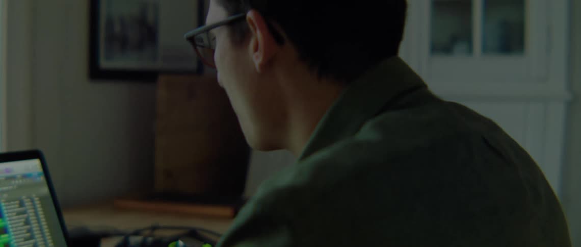 A man in a room with glasses sitting at a desk using a laptop with a window and a wall in the background