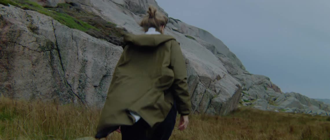 A woman wearing a coat is walking through tall grass near a mountain to a building in the distance