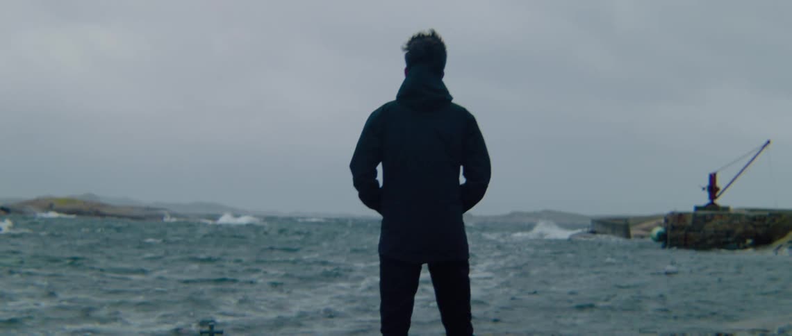 Man with glasses black coat and pants stands and watches sea on cloudy day