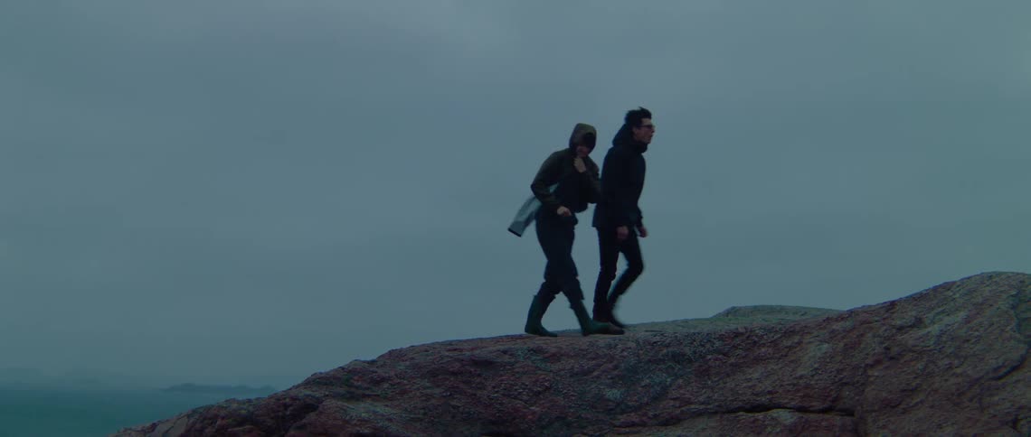 Man and woman with glasses raincoats pants and boots walk on the side of cliff on a cloudy day