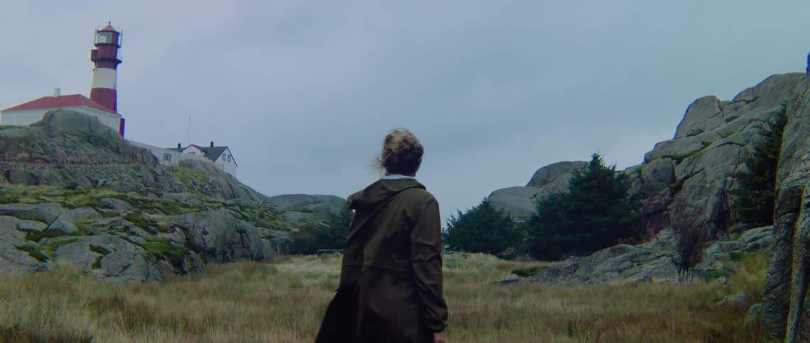A woman is standing outside with the wind blowing her hair as she looks around at rocks and there is a lighthouse in the distance