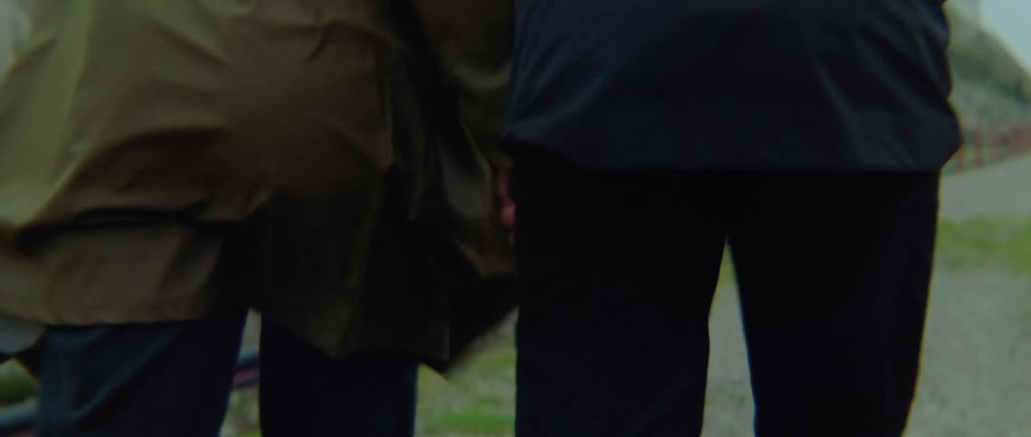 A man and a woman holding hands are walking along a gravel driveway in a coastal region on a windy day