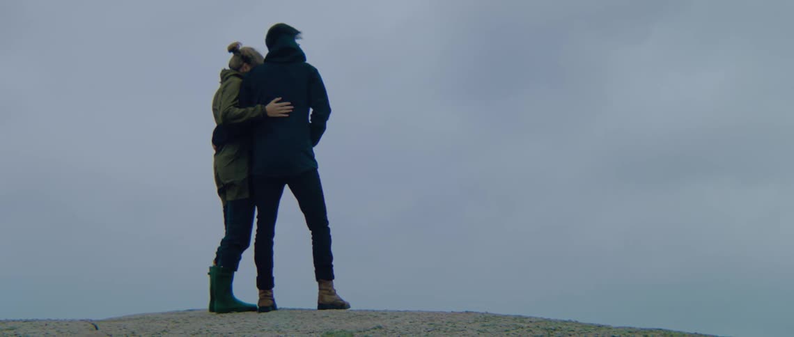 A man and woman stand with arms around each other on a ridge rock on a windy day