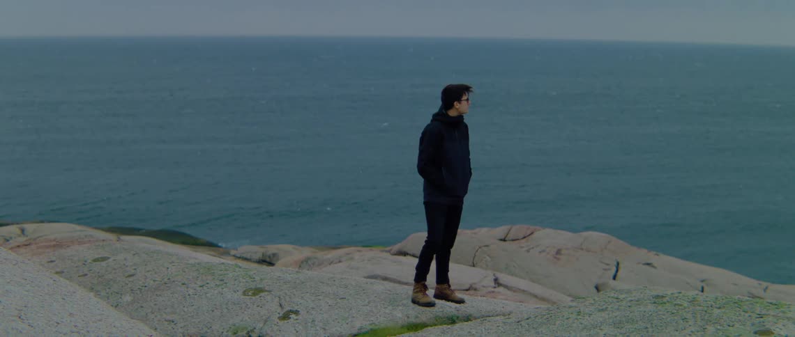 A person standing on a rocky shore with the ocean sky and clouds in the background