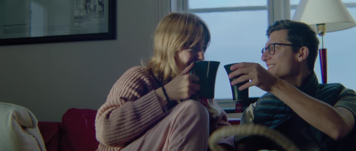 Man with glasses black vest and t shirt and blonde woman in pink sweater and pants drink two cups of tea in living room at home