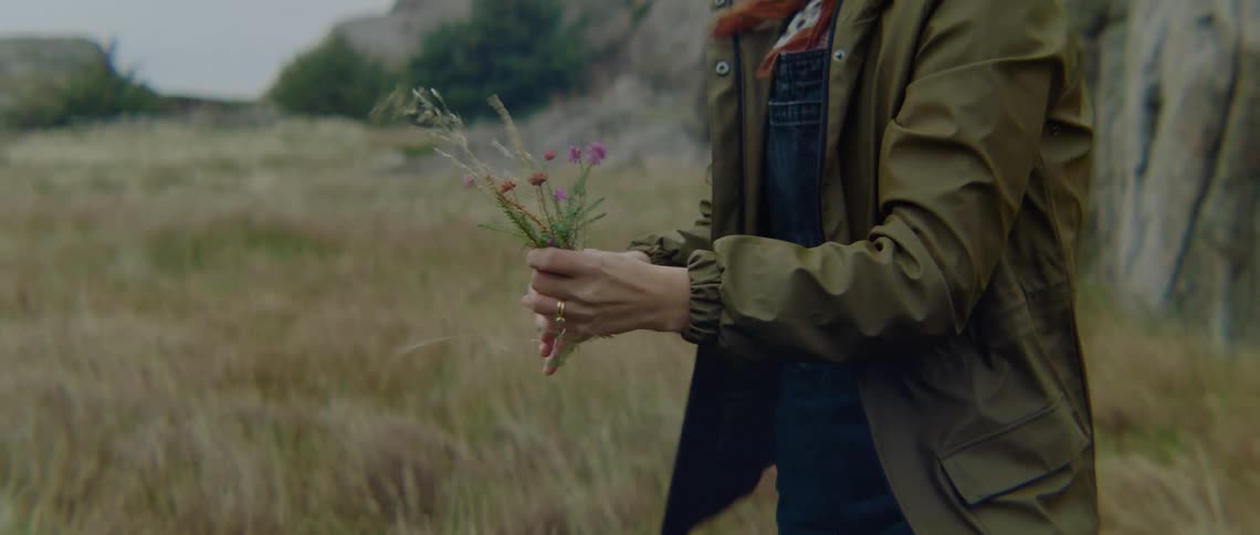 Woman with brown coat green boots and tied kerchief pick up wild flowers in the field
