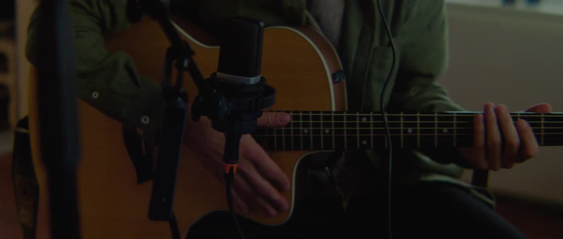 A man in a room is sitting and playing a guitar while wearing headphones and recording equipment is in use