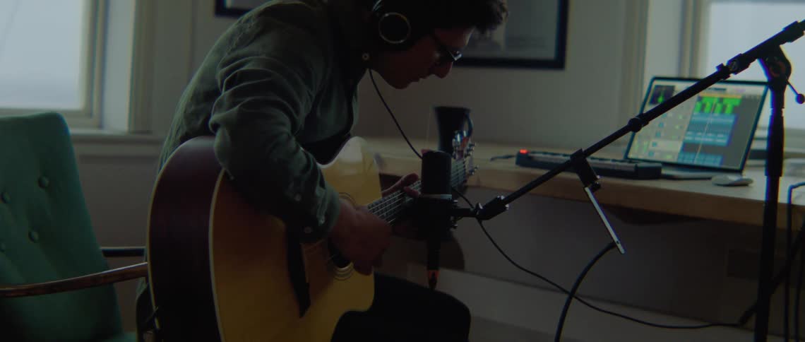 A man with glasses playing a guitar in front of a microphone and using a laptop
