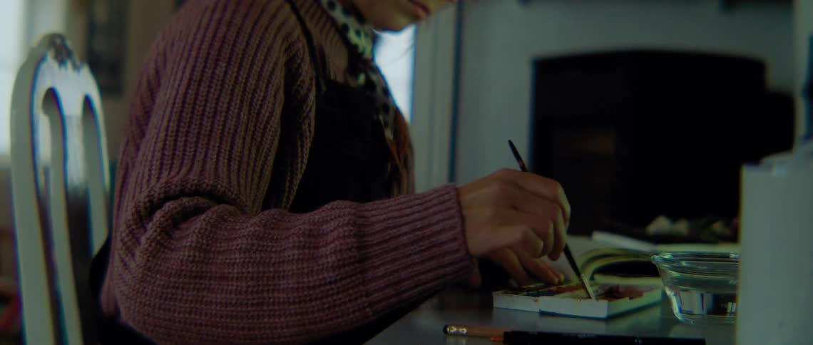 A woman sitting on a chair at a desk mixing paint with a paintbrush