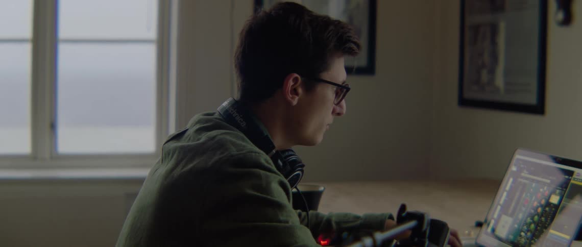 A man wearing glasses is sitting in front of a laptop computer with headphones around his neck as he works