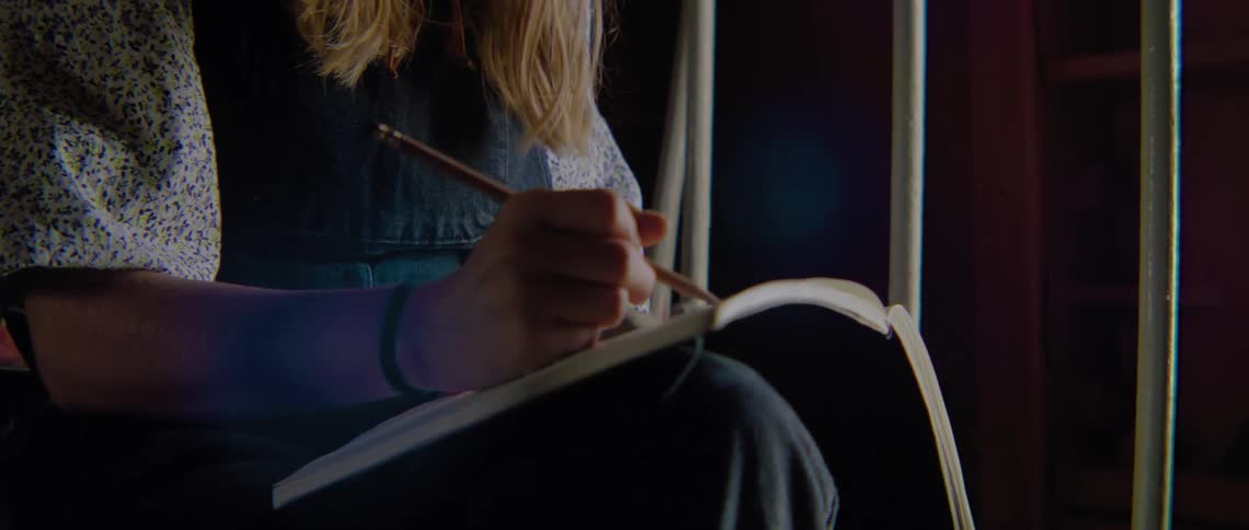 A girl is sitting in a room and writing in a notebook with a pencil