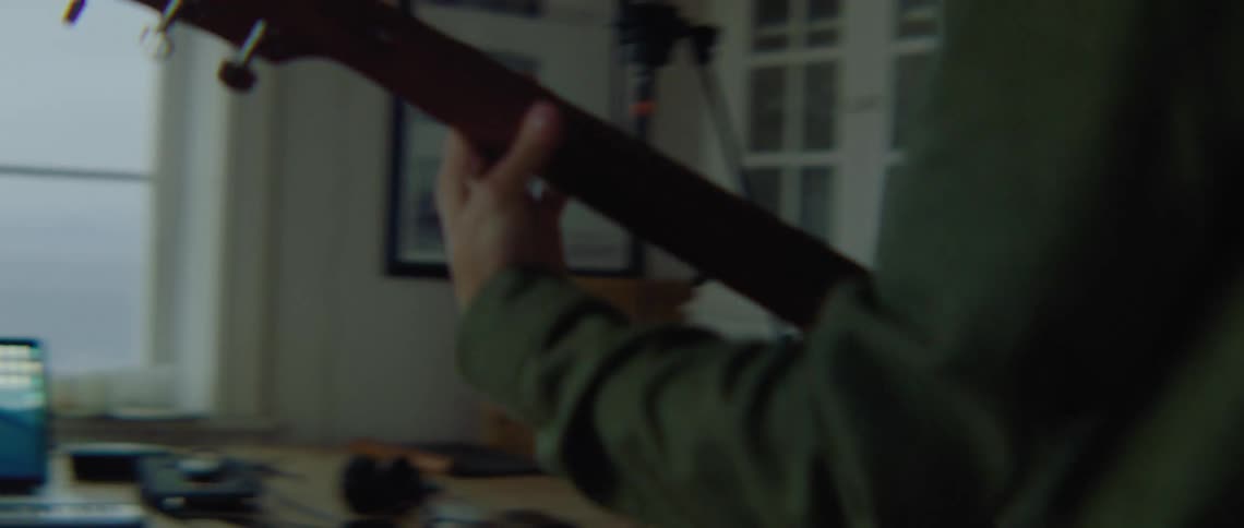 A man with glasses and a guitar in a room using a laptop on a table with a wall in the background