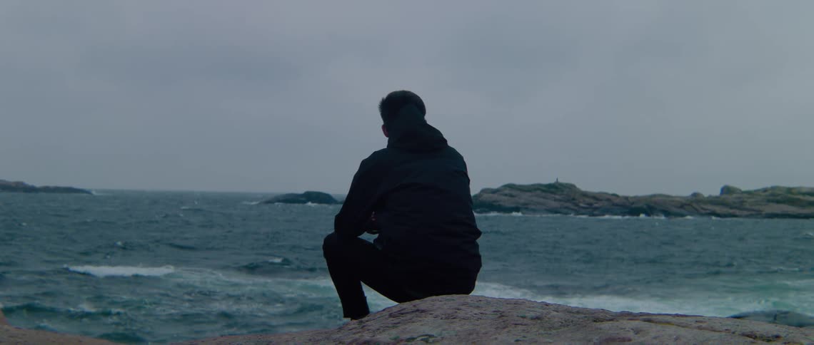 A man sits on the rocky shore looking out at the ocean as waves splash up