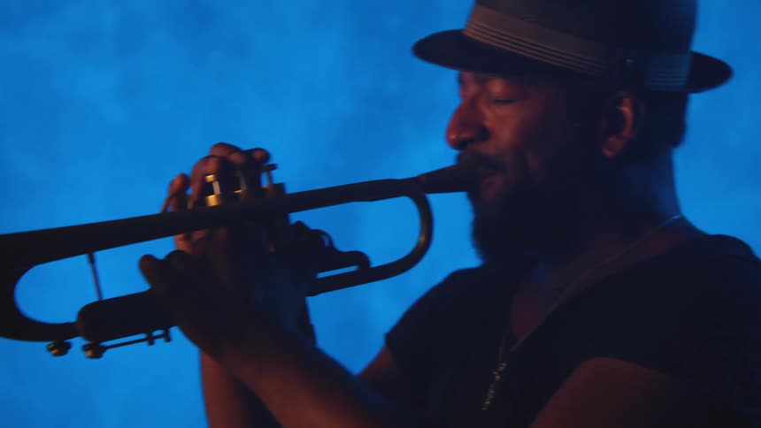 A man with a hat playing a trumpet with a wall and a screen in the background