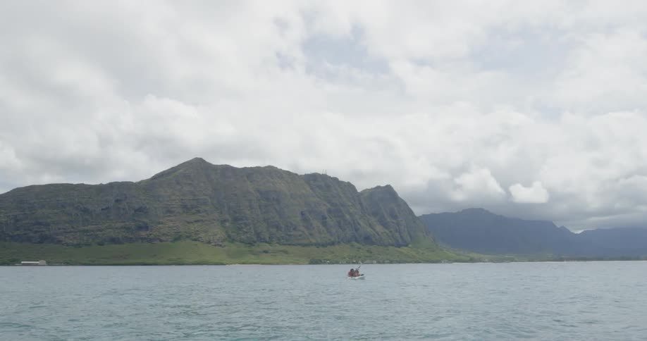 People rowing a small boat in a large body of water with mountains in the background on a cloudy day