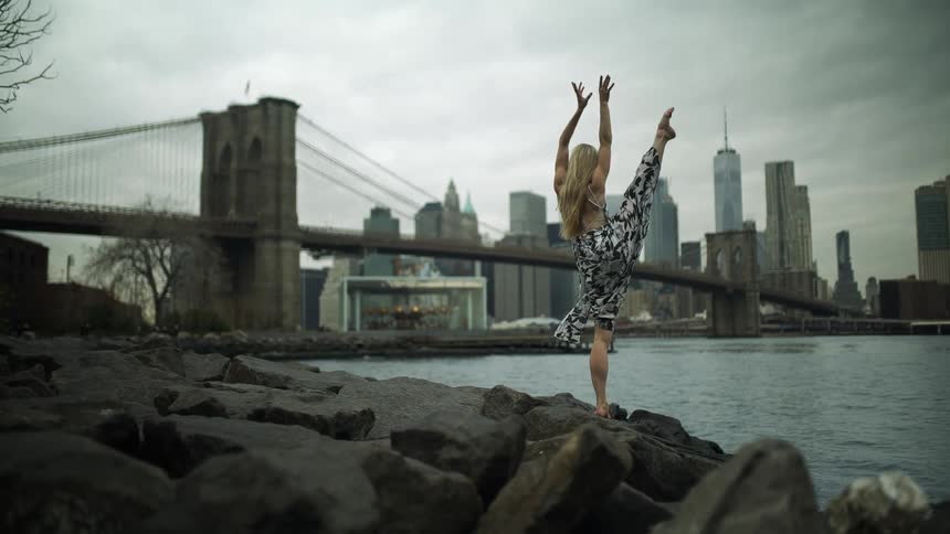A woman wearing a dress is doing yoga on large rocks next to a river with a skyline in the background on an overcast day