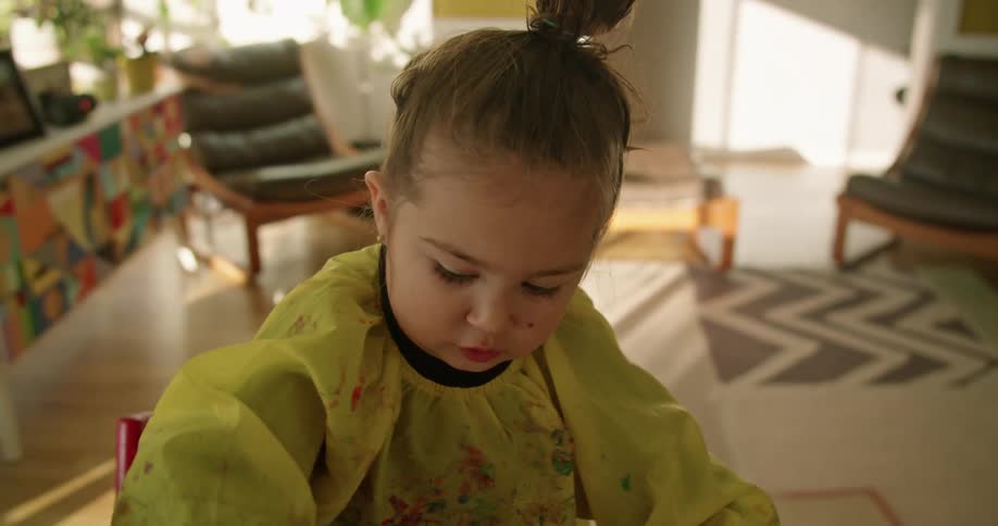 A little girl in a room painting on a paper with a wall and window in the background