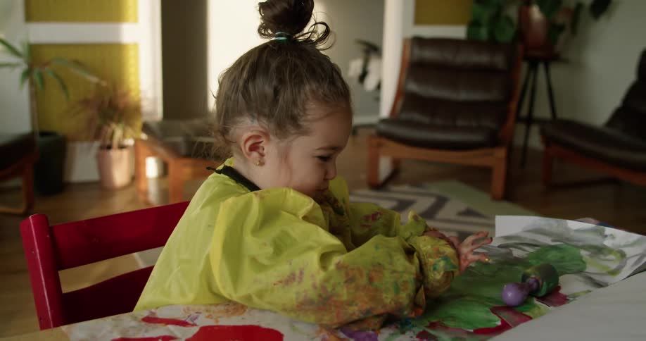A little girl is finger painting and there is paint all over her clothes