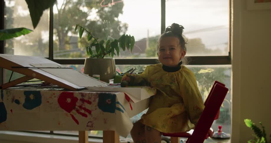 A young girl smiles as she sits at a table at home where she is painting