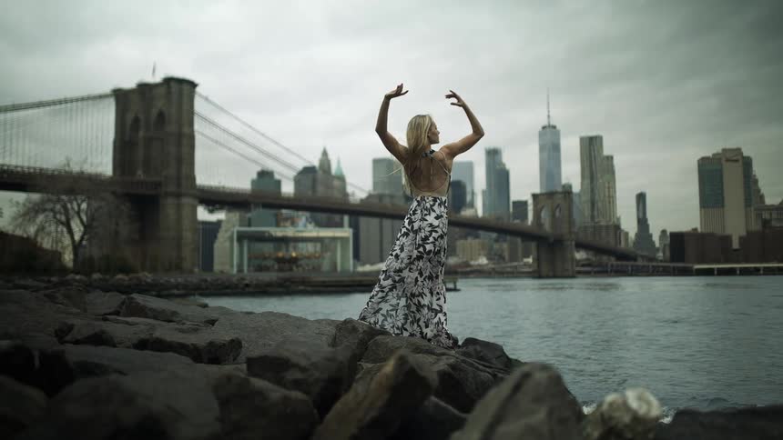 A woman is standing on the rocks of a riverbank and posing with a suspension bridge and skyline of a large city in the background on an overcast day
