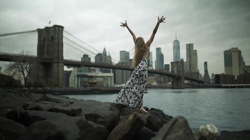 A woman wearing a black and white dress is standing on rocks along the side of a river and stretching her arms out in the air