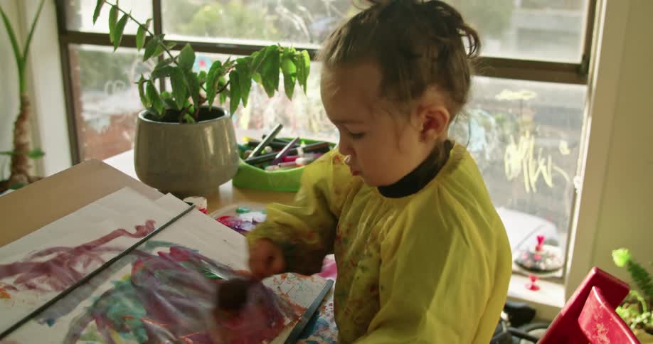 A little girl in a room painting on a paper with a wall and window in the background