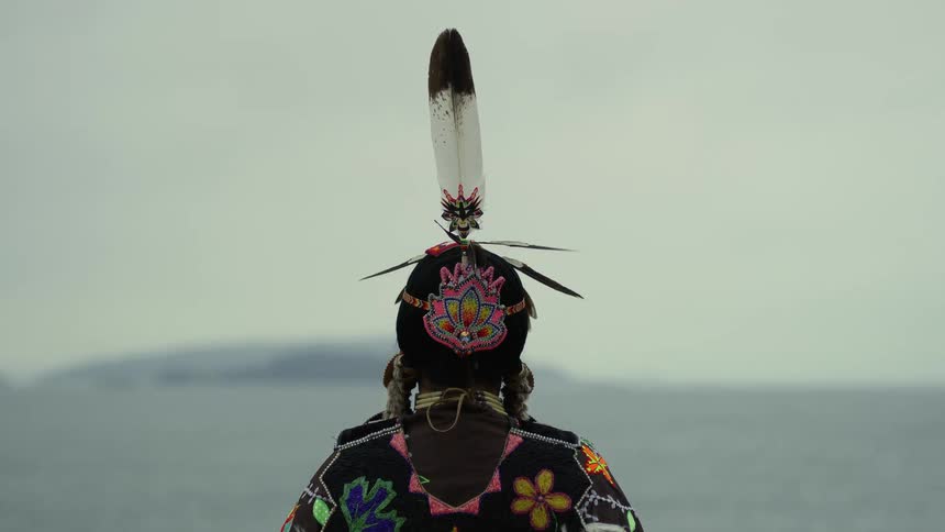 A native american indian with one tall feather on his head is looking at the water