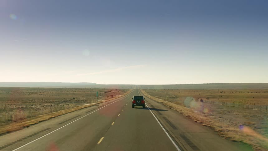 A car travels on a desolate stretch of road with the sun shining and glaring