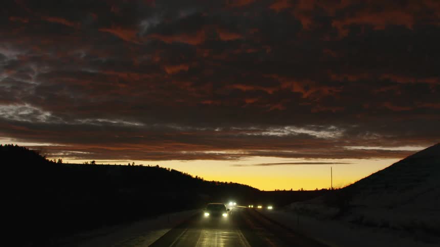 Cars are traveling on a road with the headlights on under a colorful cloudy sky