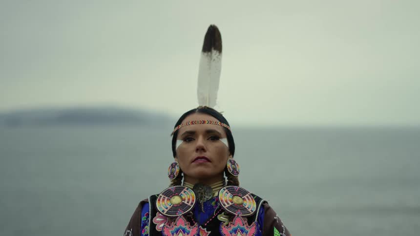 A native woman stands by the ocean in traditional clothing and headdress
