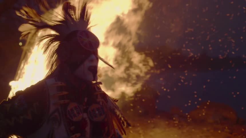 A man is dancing in front of a bonfire at night while wearing traditional native american clothing