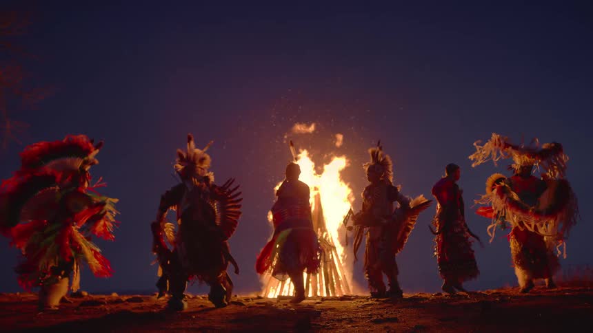 Indigenous men in traditional clothing dance around a fire