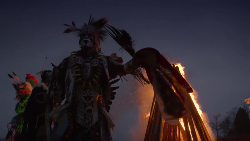 Men dressed in feathers dancing in front of a fire with trees the sky and clouds in the background