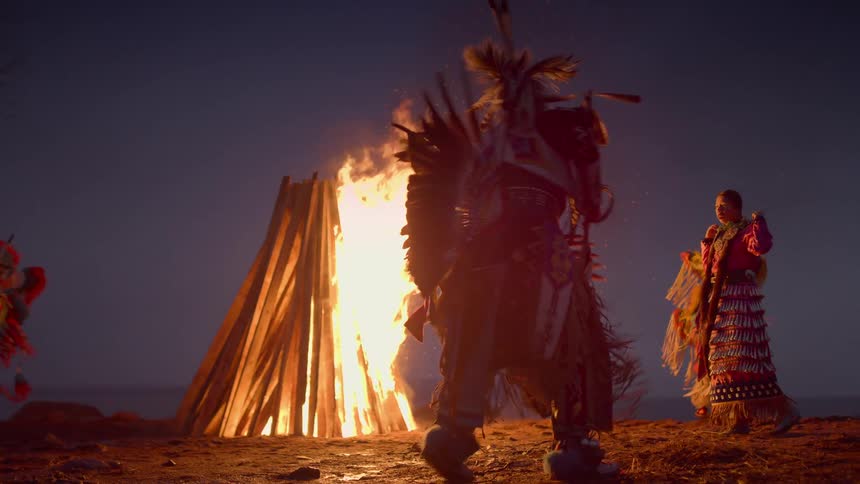 People in traditional costume dance perform traditional dance around bonfire at night with mountains on horizon