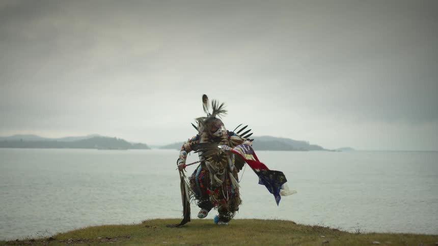 A native american powwow dancer is dancing on the shore with the water behind him