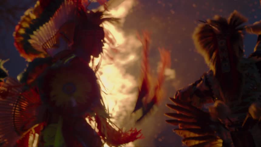 Two native americans wearing traditional garb dance outside at night around bonfire