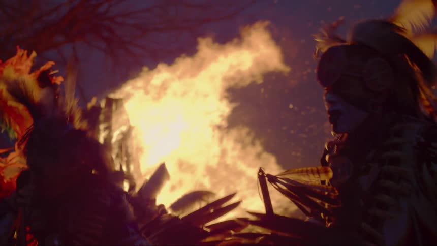 Men wearing traditional amerindian headdress and garb dance near bonfire at night near leafless tree