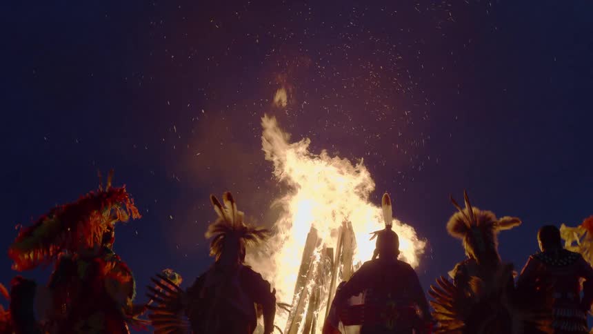 Indigenous people are dancing around a bonfire at night while dressed in traditional clothing