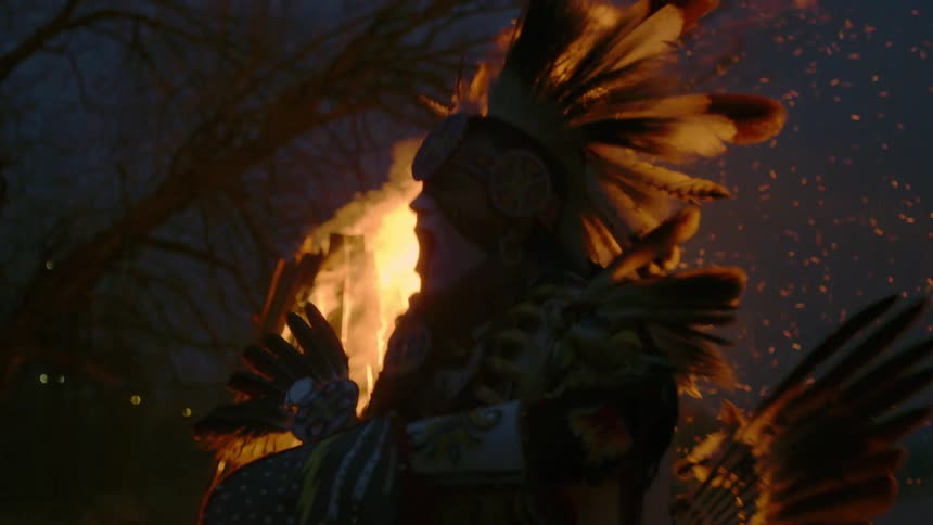A man dressed in feathers dancing in front of a fire with trees the sky and clouds in the background