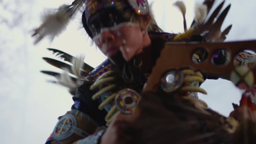 A man is performing an native American dance while dressed in traditional clothing and accessories during the day