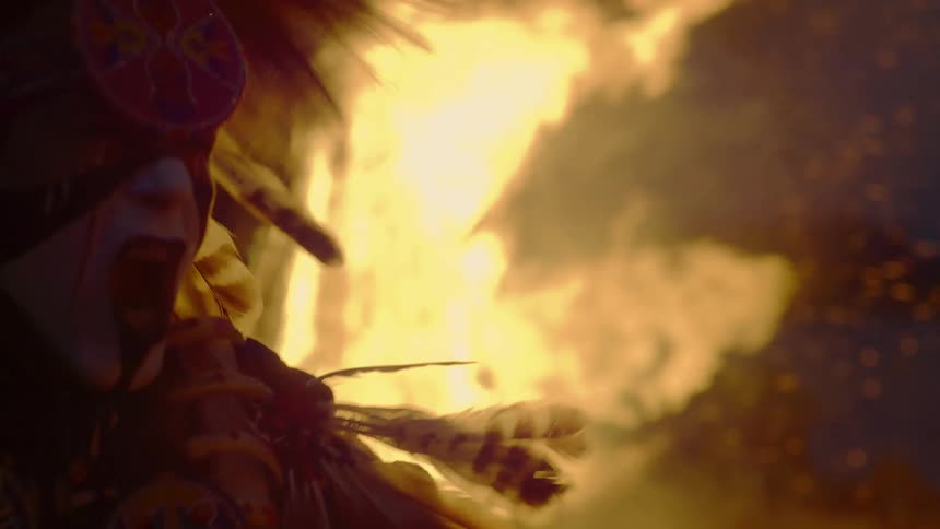 A native american with a feather headdress dancing in front of a large fire at night