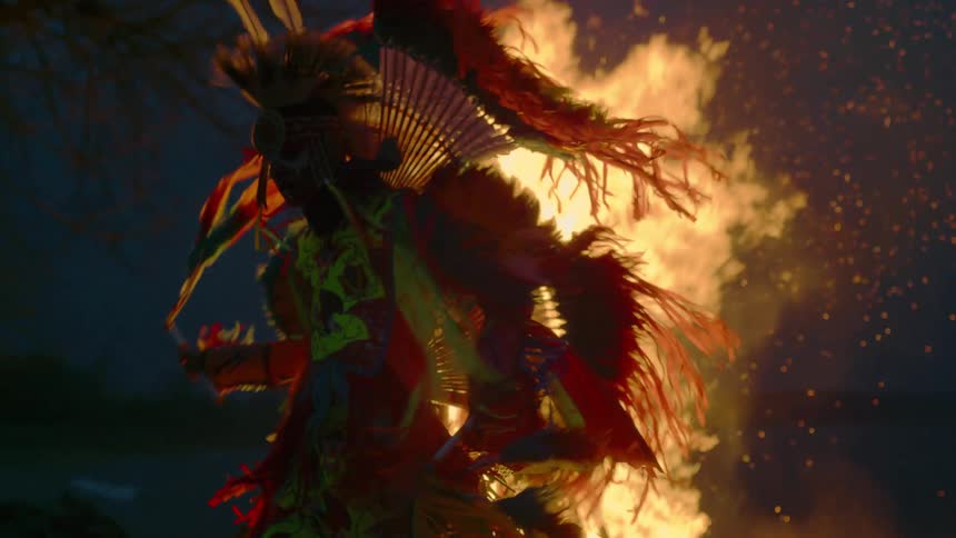 A native american pow wow dancer is dancing as a fire burns behind him and hot sparks fill the air