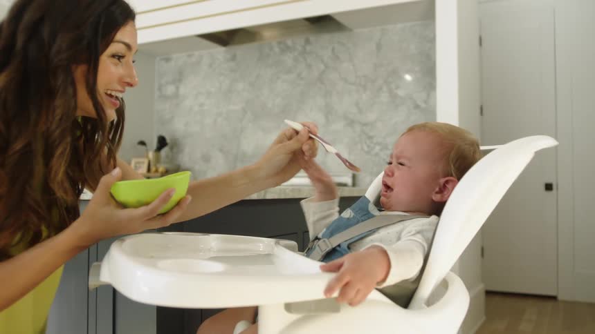 A mother trying to spoonfeed her crying baby in a highchair
