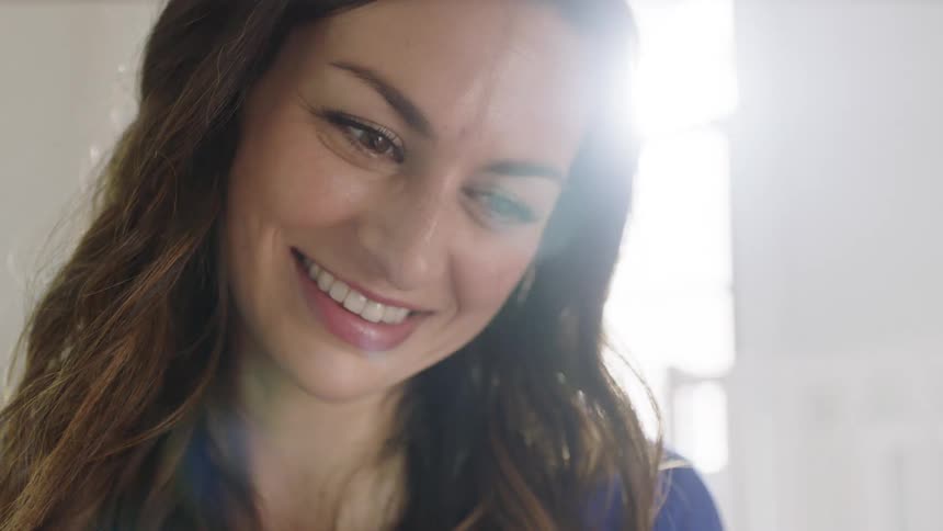 A woman is looking down and smiling in a room in front of a sunny window