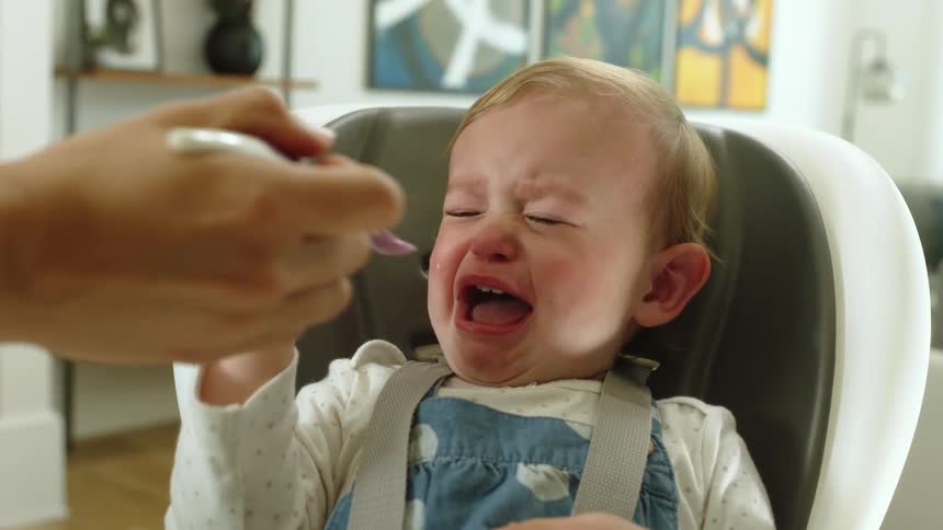 A baby in a high chair pushes away a spoonful of food and cries