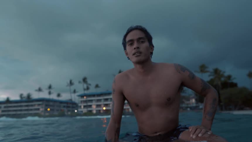 A man on a surfboard in the ocean with buildings trees the sky and clouds in the background