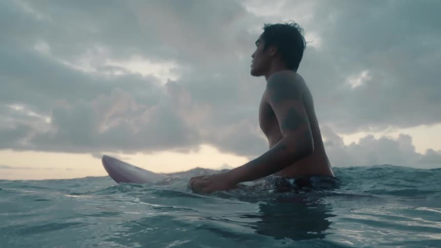 A male surfer sits atop his surfboard while waiting for his next wave to ride