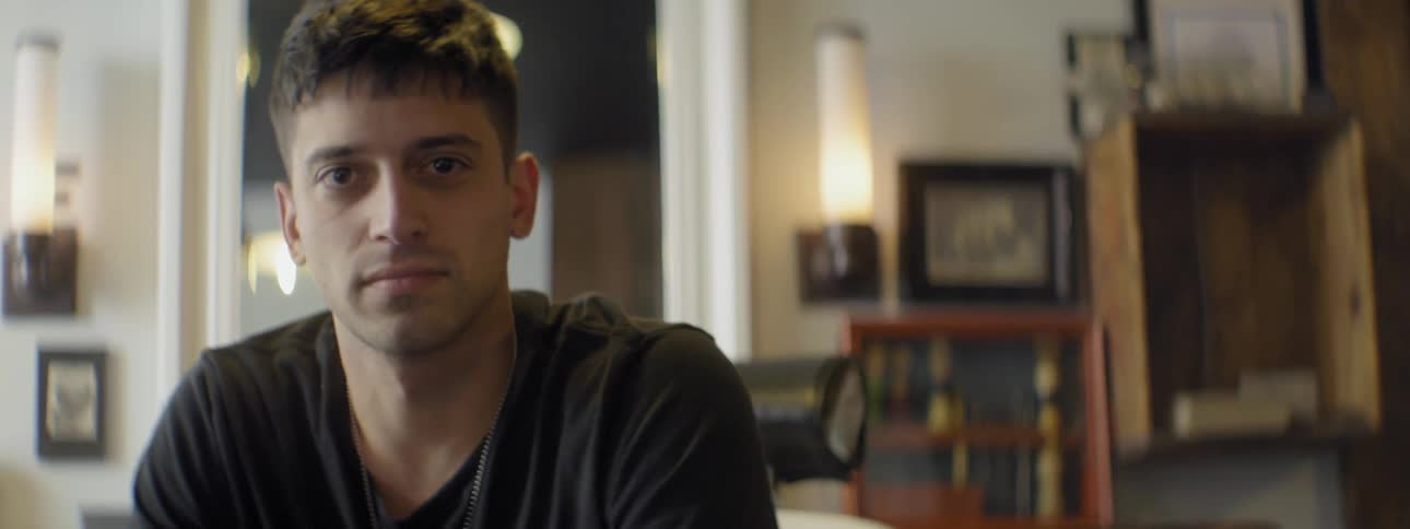 A young man sitting in a room turns and looks directly at the camera